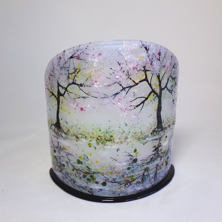 Curved glass sculpture on a black base. Two trees in blossom are reflected in water