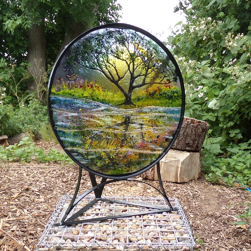 Circular glass sculpture with a painted metal frame. The design is an arching oak tree at sunset. The sculpture is photographed in a woodland with sunlight shining through the glass.