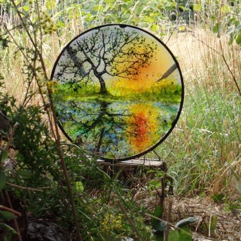 Glass disc with a design of a tree reflected in the canal. Framed in black metal. Photographed in woodland.