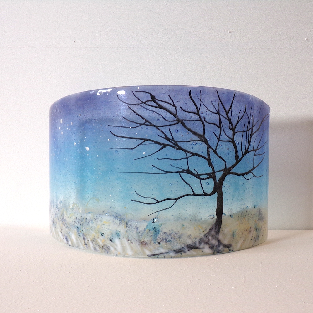 Curved glass sculpture featuring a tree in a snowy landscape with a cool blue night sky