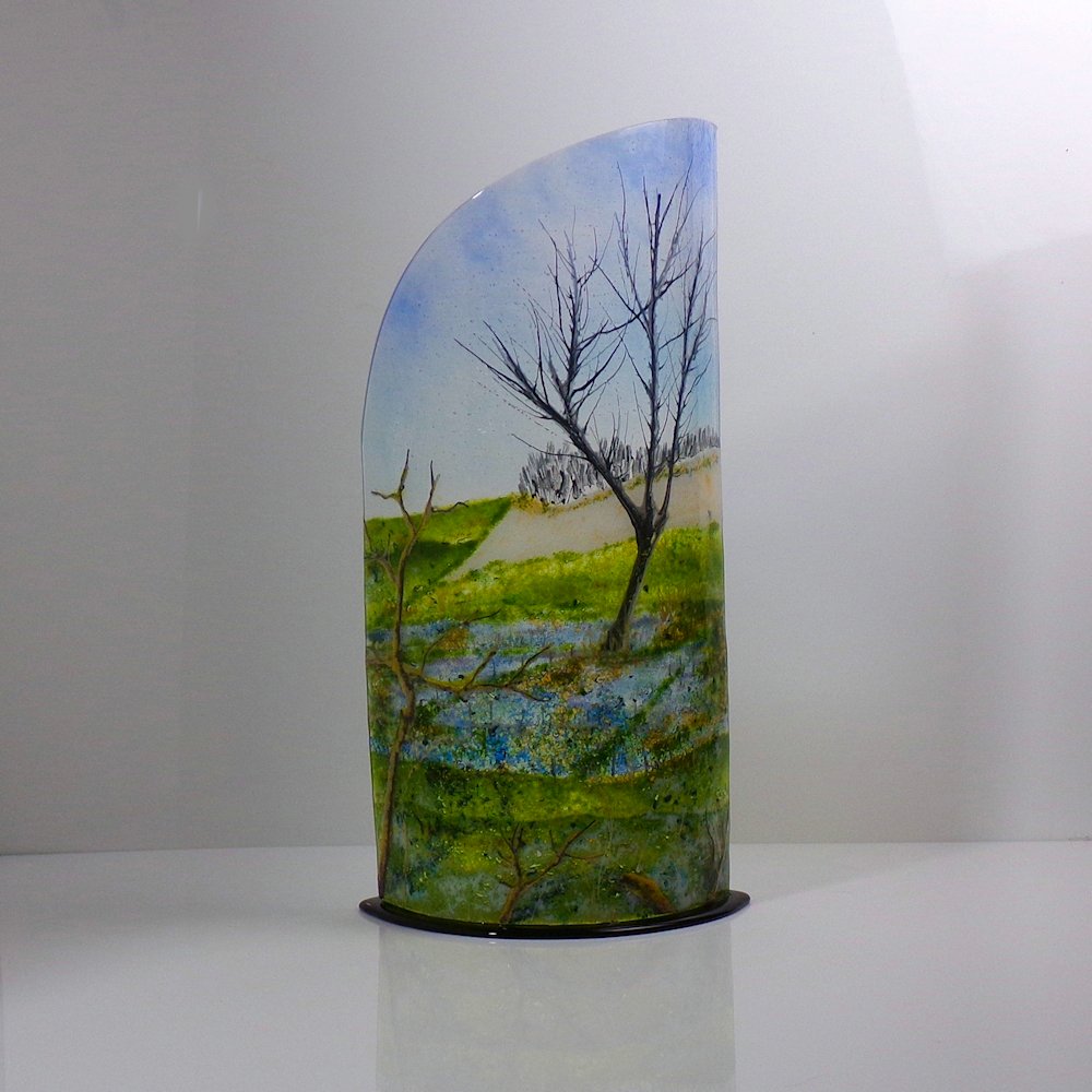 Curved glass sculpture on a black glass base showing a tree with water and snow in the background.