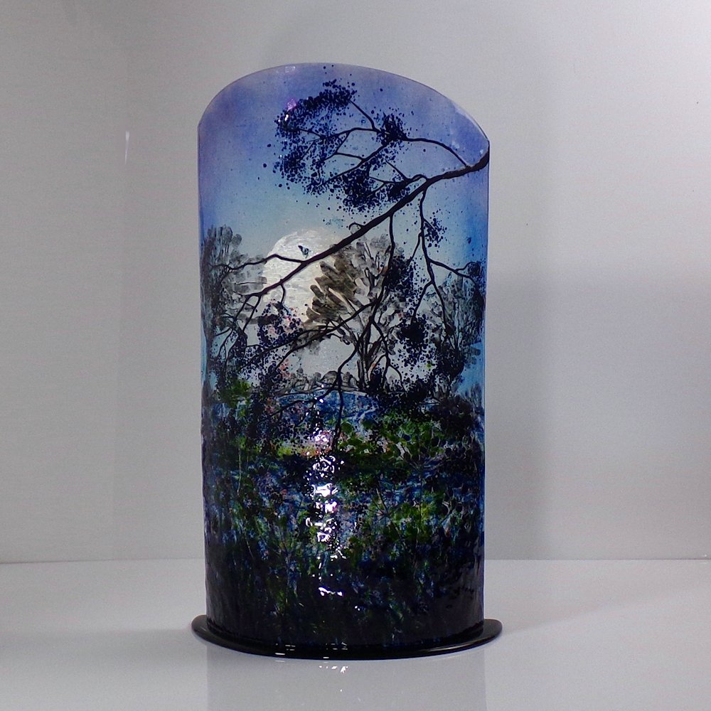 Large curved glass sculpture on a black glass base. The moon rising behind trees.
