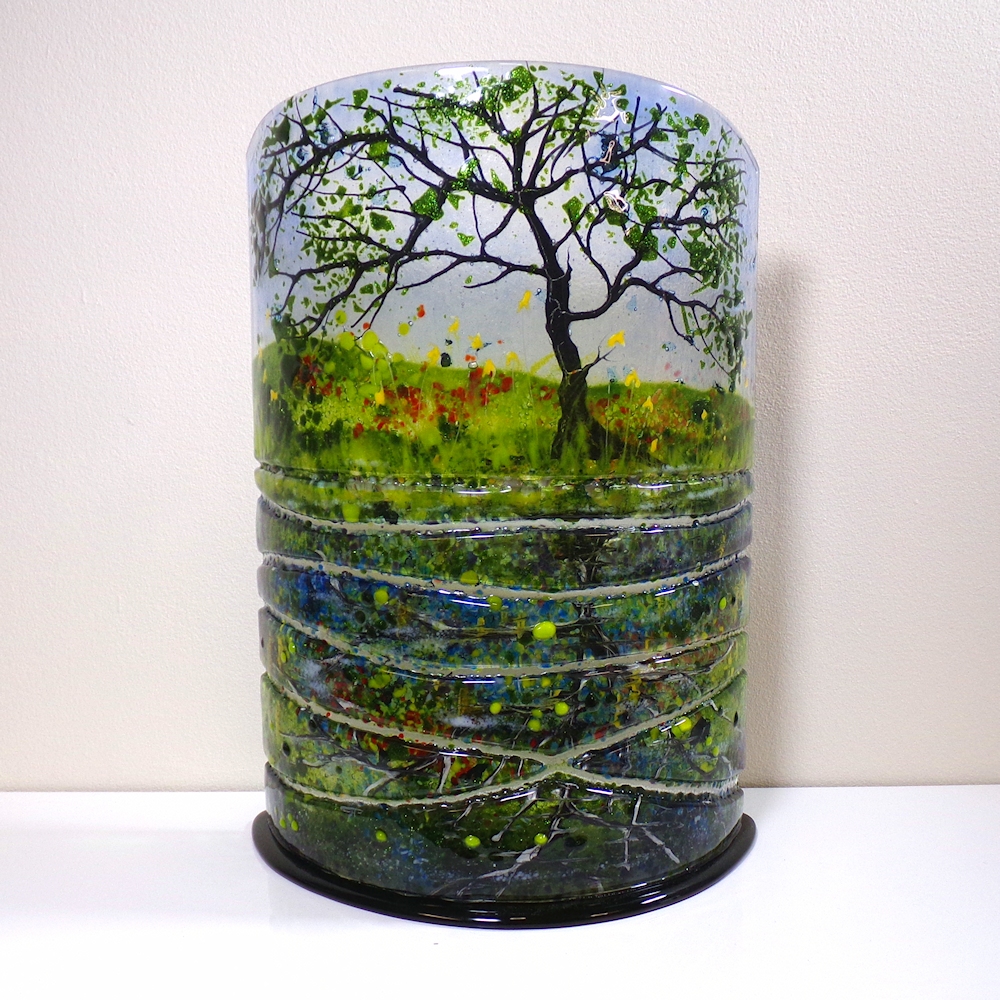 Curved fused glass sculpture, with a black glass base. Tree in the foreground with spring flowers. Reflections in the water represented by sections of glass.