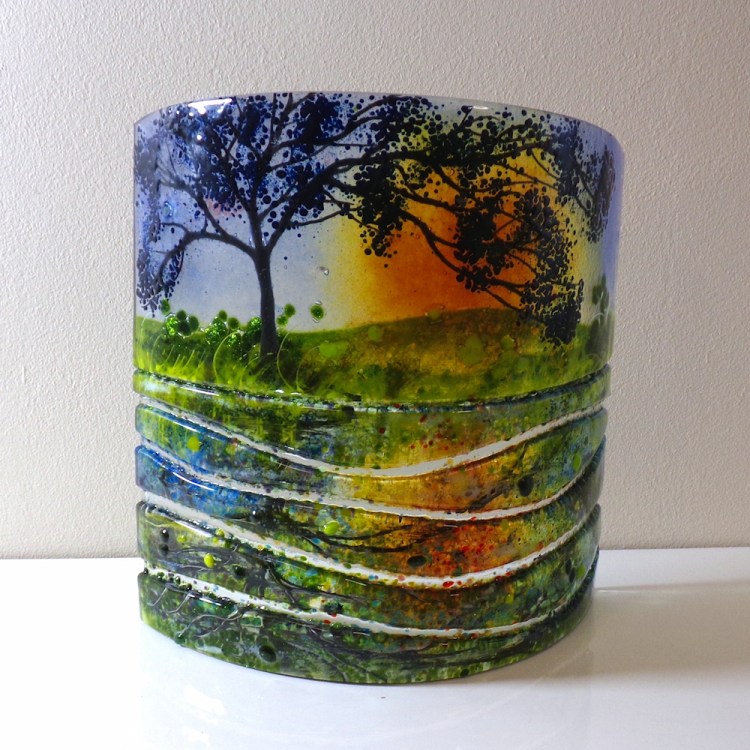 Curved fused glass sculpture, no base. Tree in the foreground with a sunset behind. Reflections in the water represented by sections of glass.
