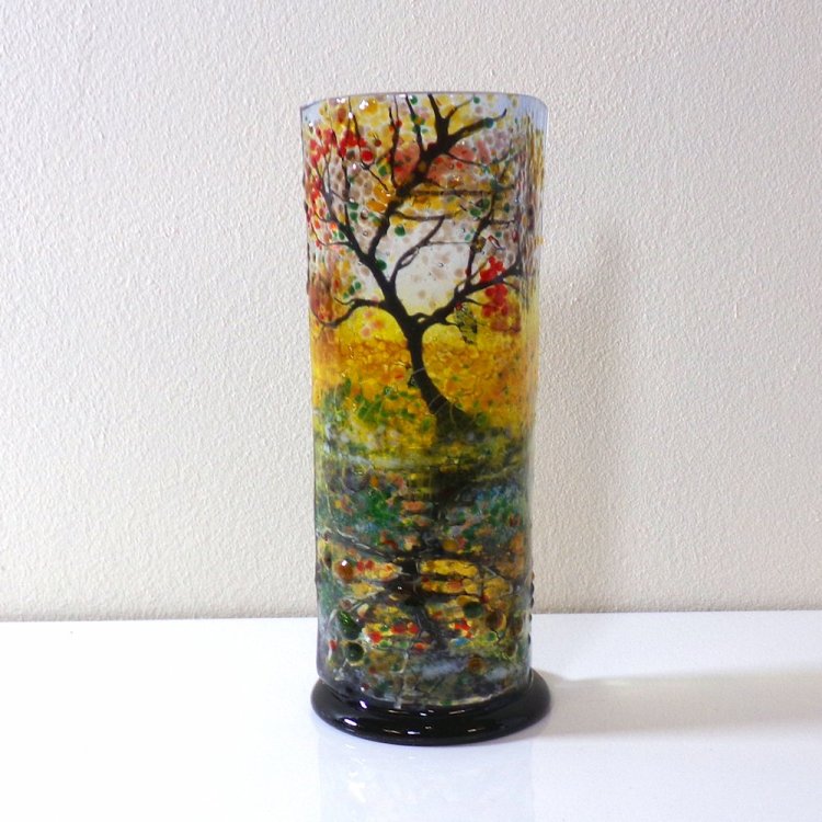 Small curved glass sculpture on a black glass base. Single tree with red/yellow leaves reflected in the canal.