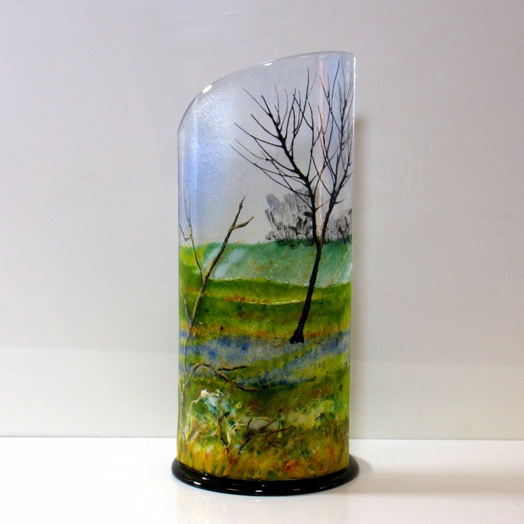 Curved glass sculpture on a black glass base showing a tree with water and snow in the background.