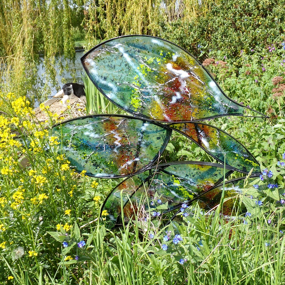 Coloured fused glass in leaf shaped sections in mild steel frame. Photographed amongst wild flowers.