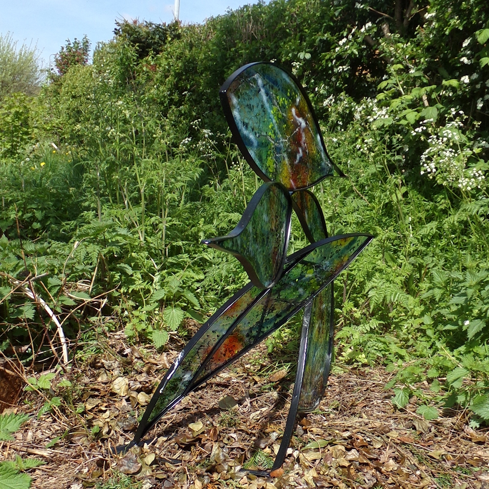 Coloured fused glass in leaf shaped sections in mild steel frame. Photographed in hedgerow.