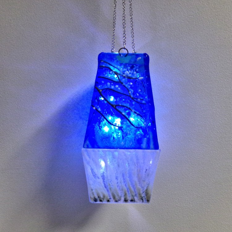 Fused glass lantern containing LED lights. Top sections show snow covered tree branches with a blanket of snow on the bottom sections.