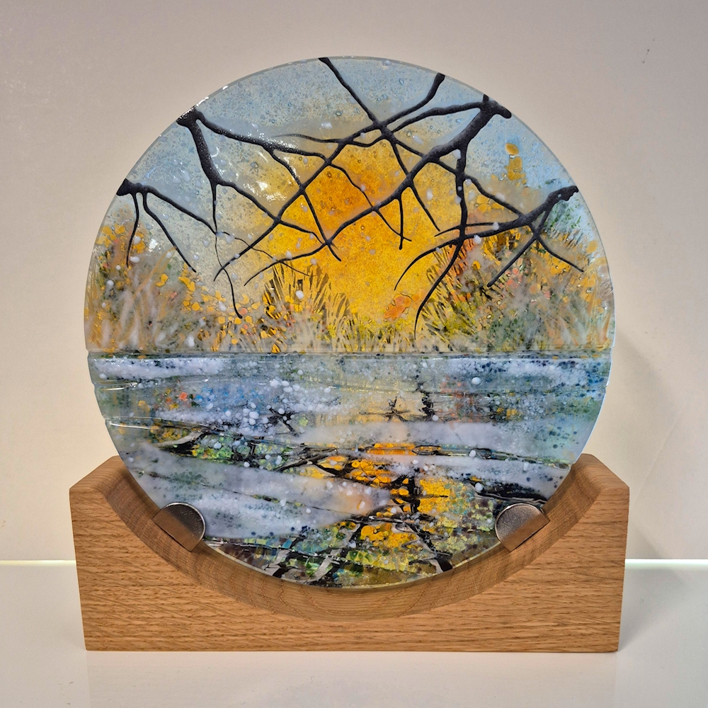 Glass disc mounted onto a wooden base. A snowy scene of trees at sunset reflected in the canal.
