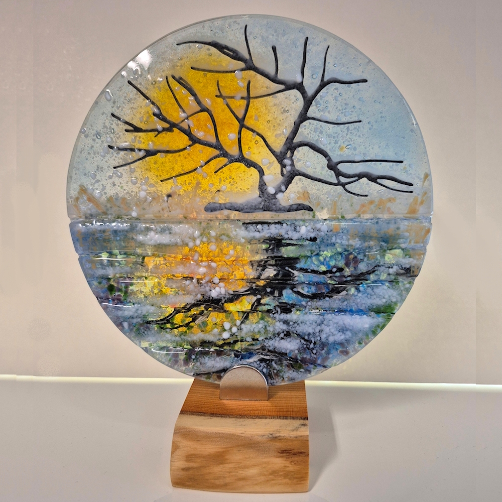 Glass disc mounted onto a wooden base. A snowy scene of a tree at sunset reflected in the canal.