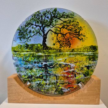 Fused glass disc mounted on a oak stand. The disc shows a tree at sunset reflected on the canal.
