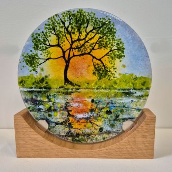 Fused glass disc on a wooden base.. A Spring scene with a tree in the foreground with the sun setting behind. The scene is reflected in the canal with textured glass representing patterns in the water.