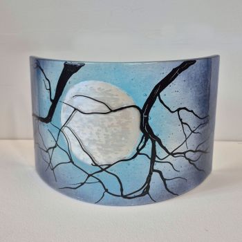 Fused curved fused glass sculpture. Two branches are in the foreground, with the moon behind them on a bright blue sky.