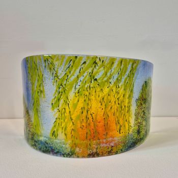 Curved fused glass sculpture with the branches of a Willow tree in the foreground; with the sun setting through the leaves.