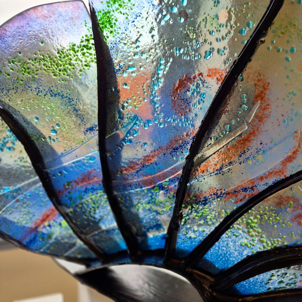 Glass and black metal sculpture. Metal is in fan shape with glass inserts between each spoke. The glass is mainly a pale blue with green and red highlights. This photo shows a close up of the central sections of glass.