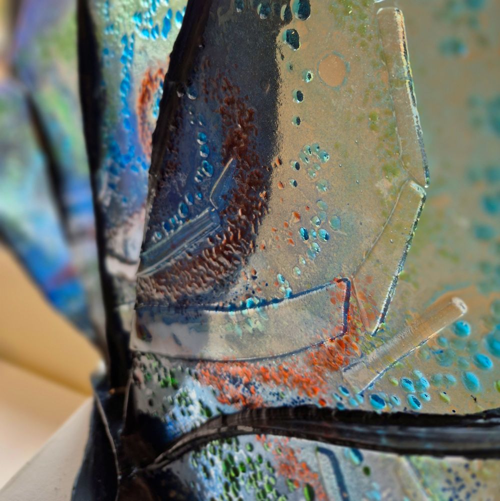 Glass and black metal sculpture. Metal is in fan shape with glass inserts between each spoke. The glass is mainly a pale blue with green and red highlights. This photo is a close up of the glass showing the texture.